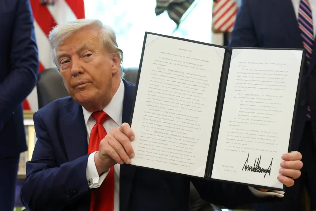 Trump assina decreto que visa acabar com políticas de fiança sem dinheiro Donald Trump segurando uma cópia de documento assinado, em evento oficial. Imagem traz foco na expressão séria do ex-presidente dos Estados Unidos e na importância do documento apresentado.