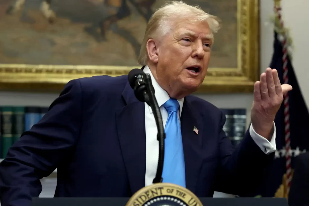 Imagem de Donald Trump falando durante uma coletiva, vestido com terno azul e gravata azul, com fundo de biblioteca e quadro ao fundo.