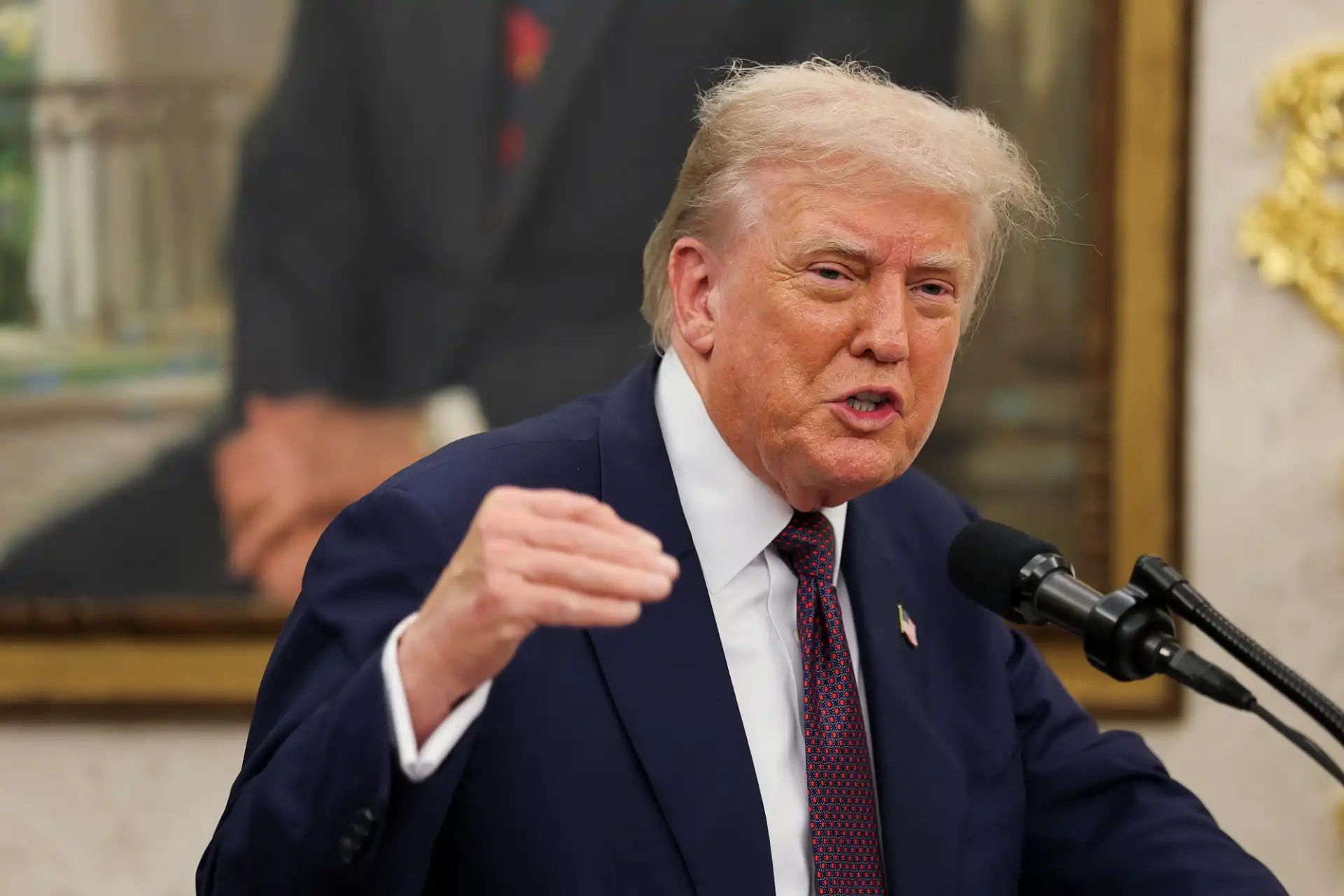 Imagem de Donald Trump falando durante uma reunião, usando terno azul e uma gravata de fundo de sala oficial com uma pintura ao fundo.