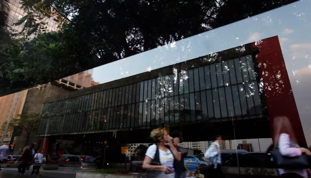Imagem de uma fachada de prédio com fachada de vidro refletindo árvores e céu, com pessoas caminhando na rua em frente, em uma área urbana moderna.