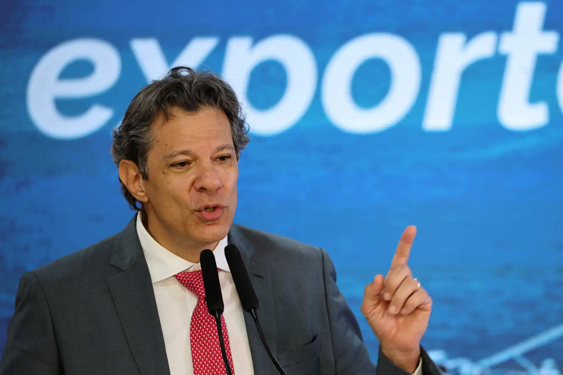 Homem falando em uma conferência ou evento com fundo azul e a palavra 'export' parcialmente visível, usando terno cinza com camisa branca e gravata vermelha, fazendo gesto de apontar com o dedo.