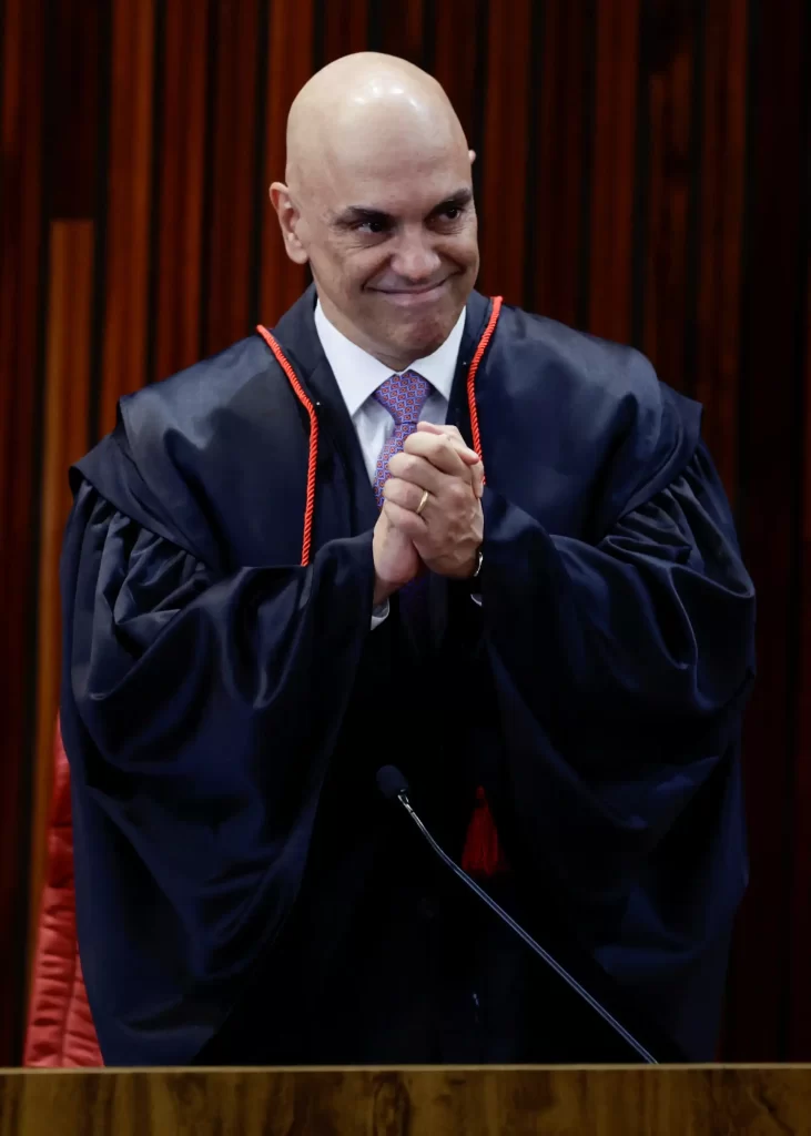 Homem vestindo toga de formatura, sorrindo e com as mãos juntas, em ambiente de cerimônia acadêmica.