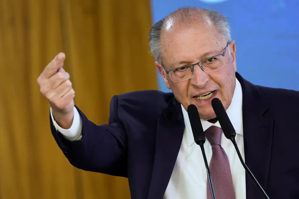 Imagem de um homem idoso com cabelos grisalhos falando e gesticulando durante uma apresentação ou discurso, usando terno e óculos.