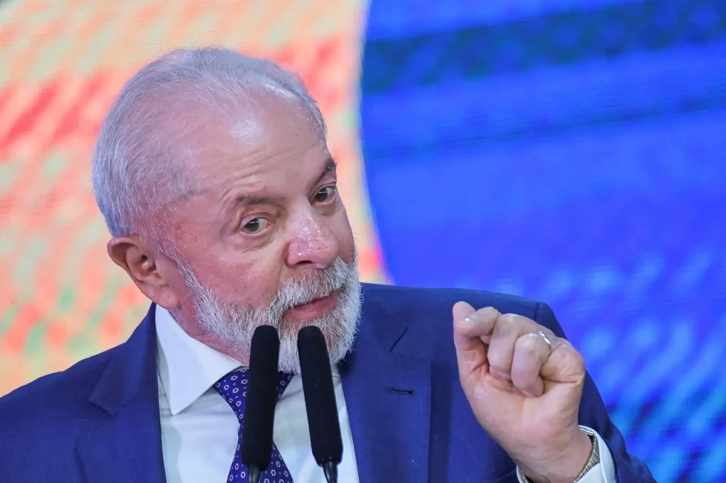 Imagem de um homem de cabelos grisalhos e barba branca, usando terno e gravata, falando em um evento com fundo colorido, representando liderança e política.