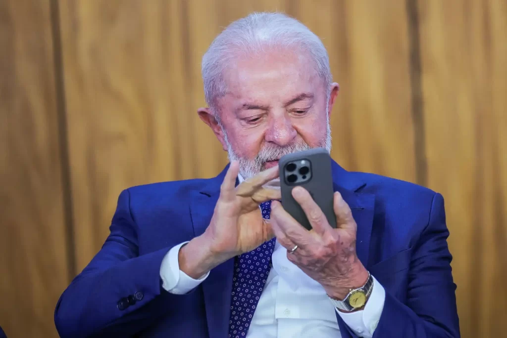 Homem idoso de cabelo branco e barba, usando terno azul, sentado em uma sala de madeira, olhando para o smartphone na mão, imagem otimizada para palavra-chave.