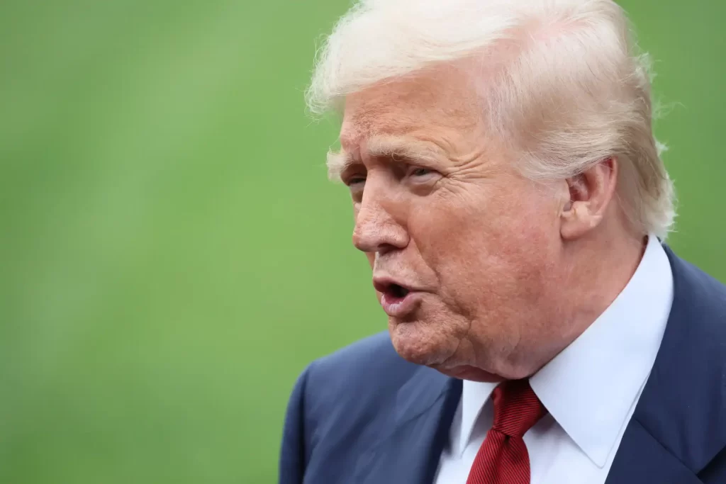 Imagem do ex-presidente Donald Trump falando, com expressão facial séria, usando terno azul e gravata vermelha, em um ambiente ao ar livre.