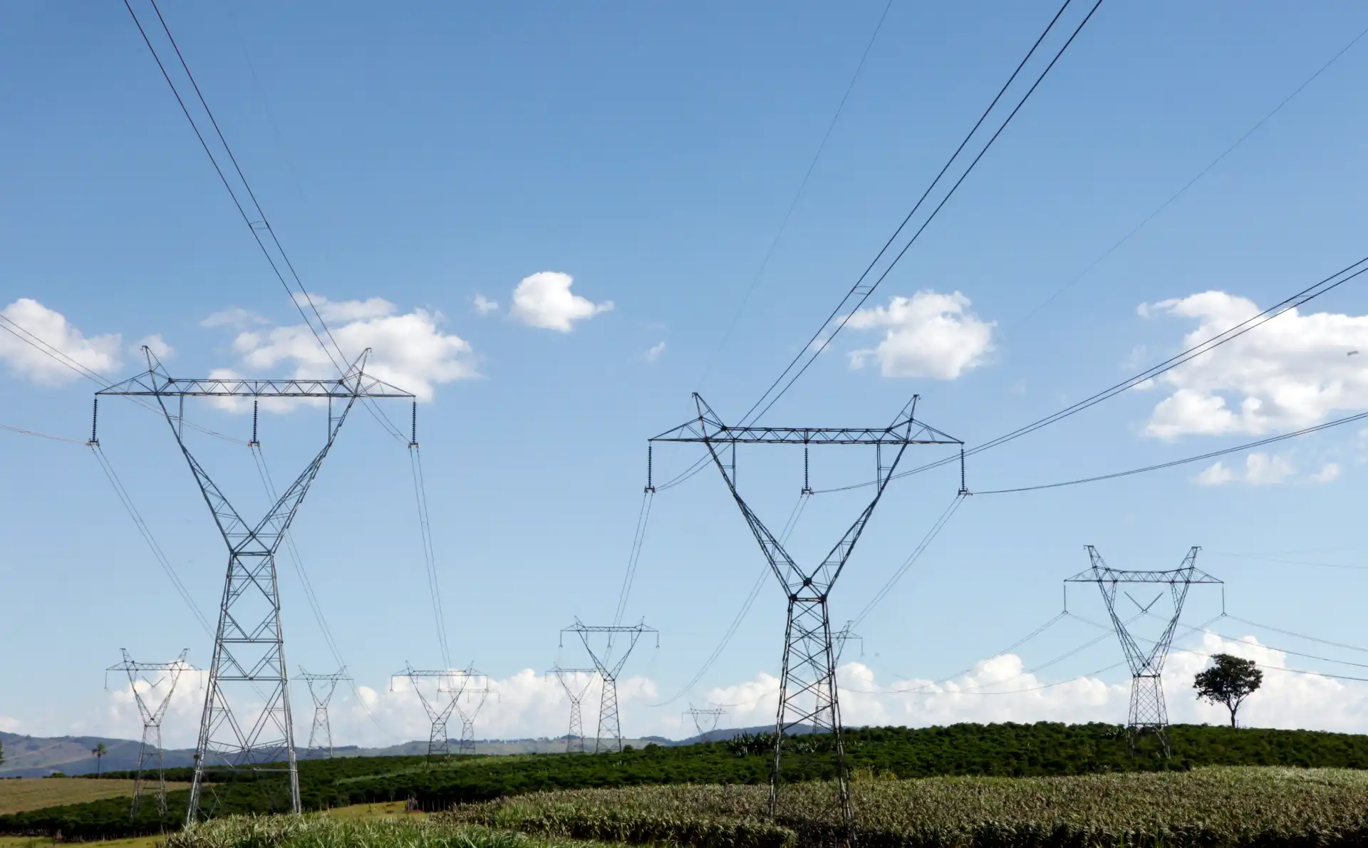 Linhas de transmissão de energia elétrica sustentando uma paisagem aberta com céu azul, nuvens brancas e vegetação ao fundo, em uma área rural.
