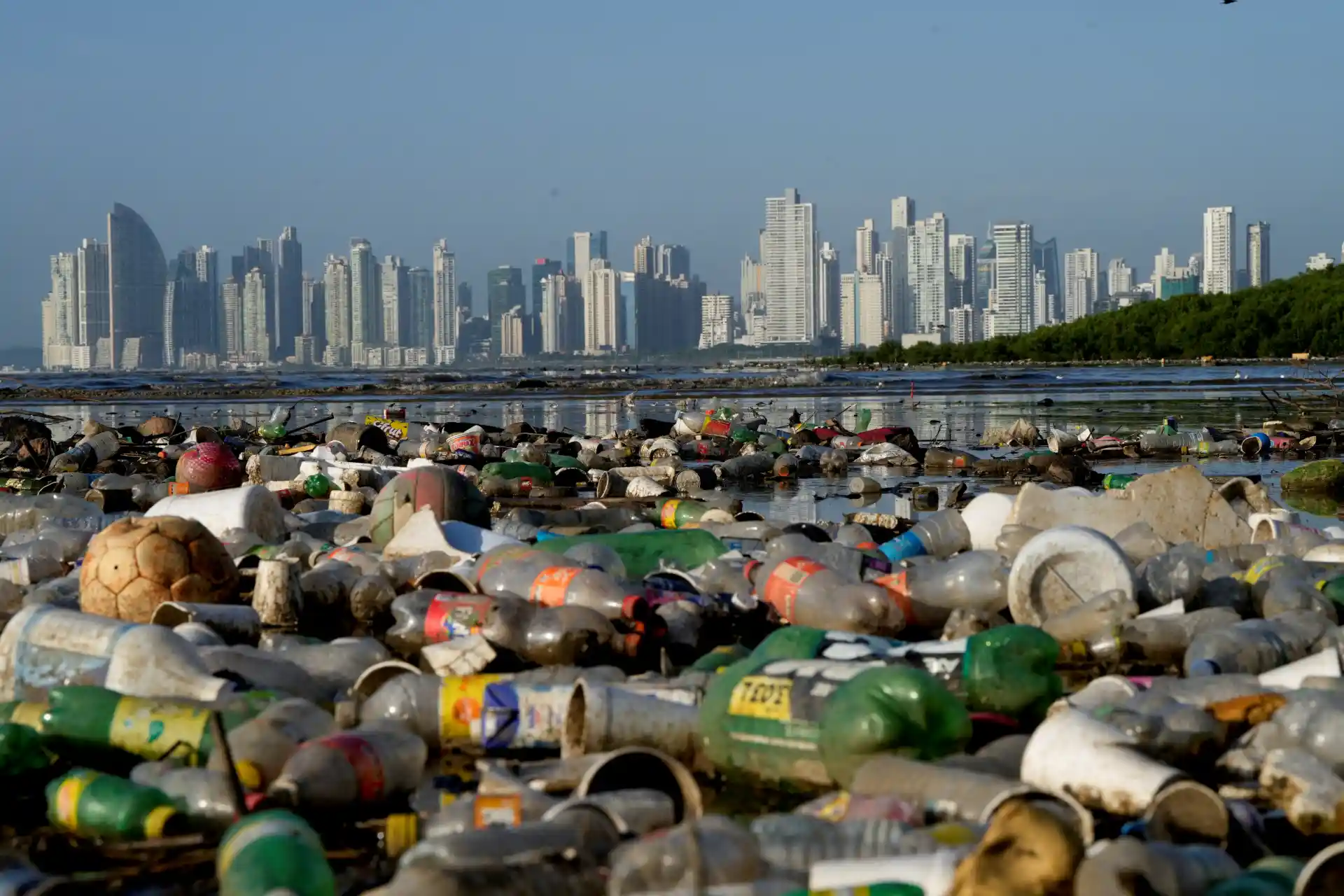 Imagem de uma grande quantidade de lixo plástico acumulado na orla de um rio ou praia com arranha-céus de uma cidade ao fundo, ilustrando a poluição ambiental e os impactos do lixo no meio urbano.