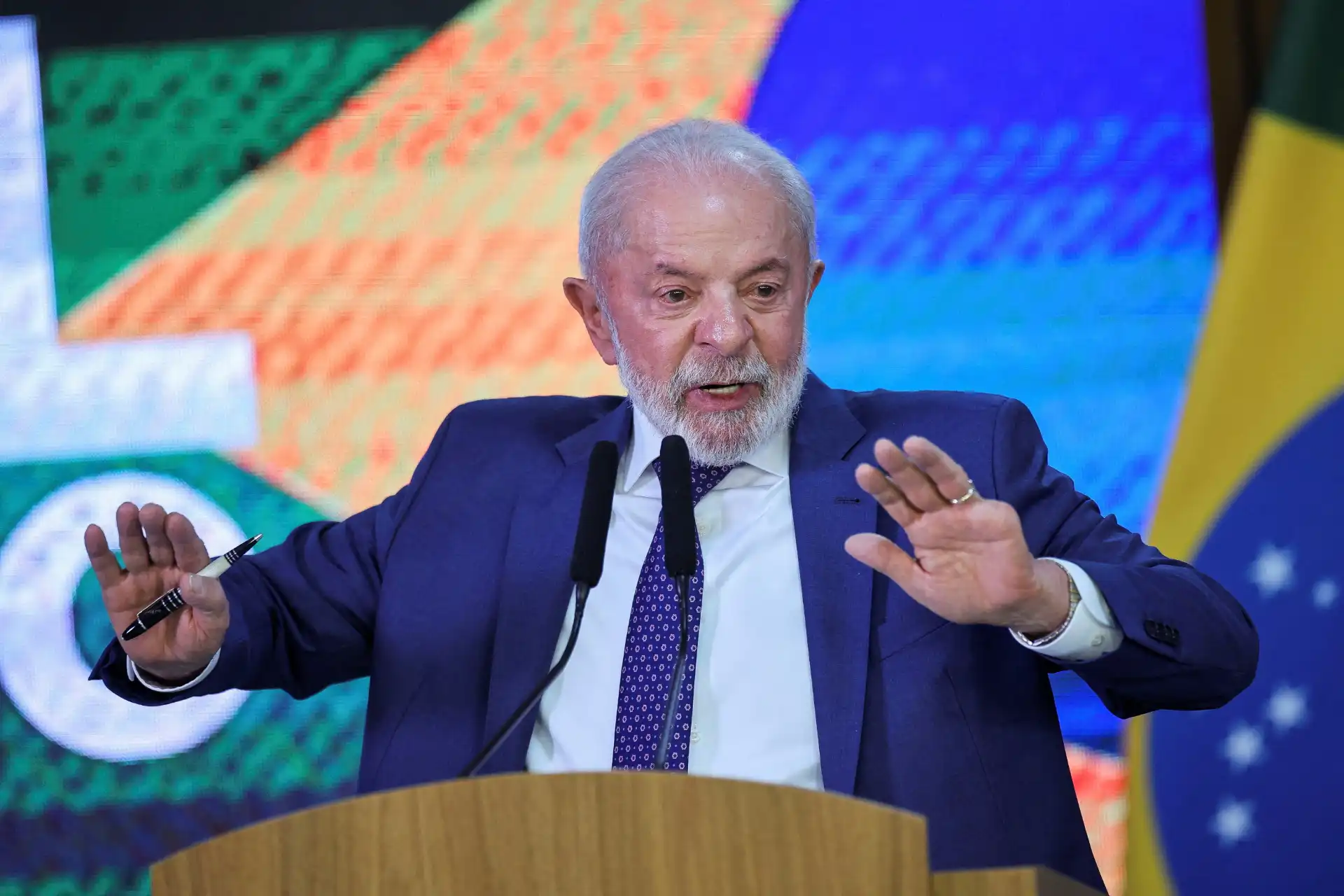 Homem idoso com cabelo branco e barba, vestido com terno azul, falando ao microfone durante uma conferência com bandeira do Brasil ao fundo.