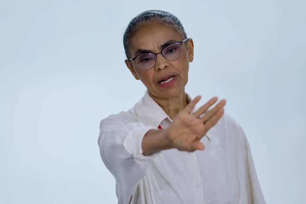 Marina Silva com óculos escuros e expressão séria, gesticulando com a mão, usando camisa branca, no fundo branco, transmitindo autoridade e assertividade.