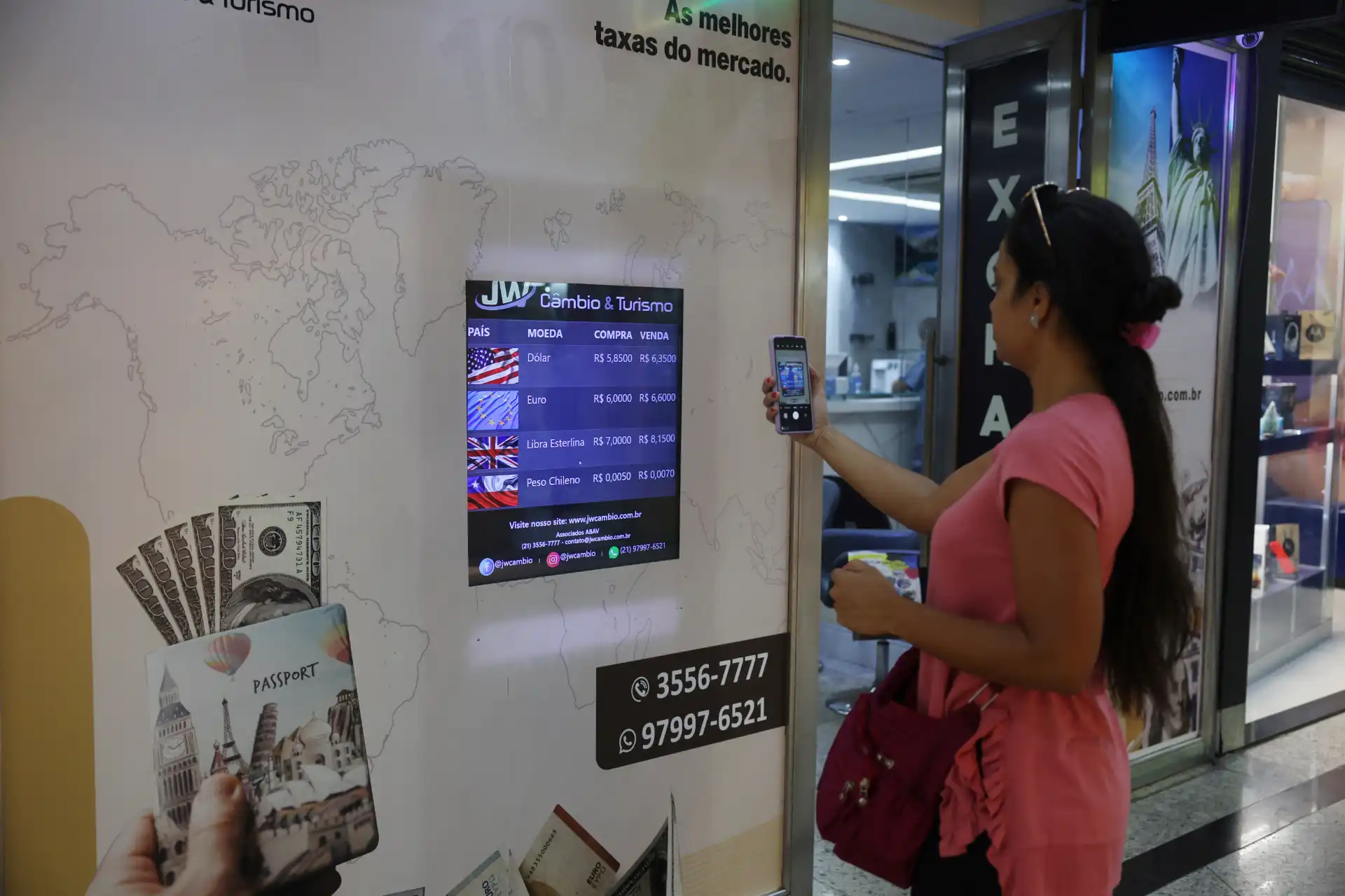 Mulher usando smartphone na exportadora de câmbio, verificando taxas de moedas estrangeiras como dólar, euro, libra esterlina e peso chileno em ambiente de loja de câmbio.
