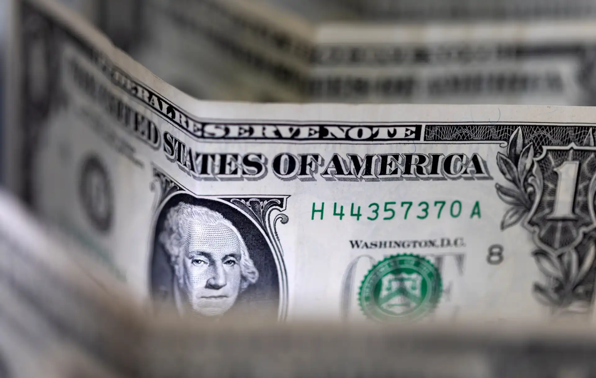 Imagem de várias notas de um dólar americano, com destaque para a nota em primeiro plano e seu número de série H44357370A, simbolizando dinheiro e economia.