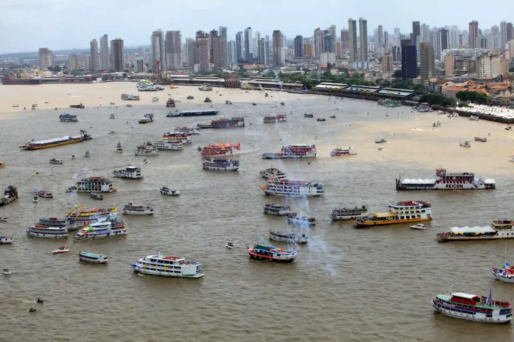 Paisagem urbana de um rio com várias embarcações, incluindo barcos turísticos e comerciais, com skyline de arranha-céus ao fundo.