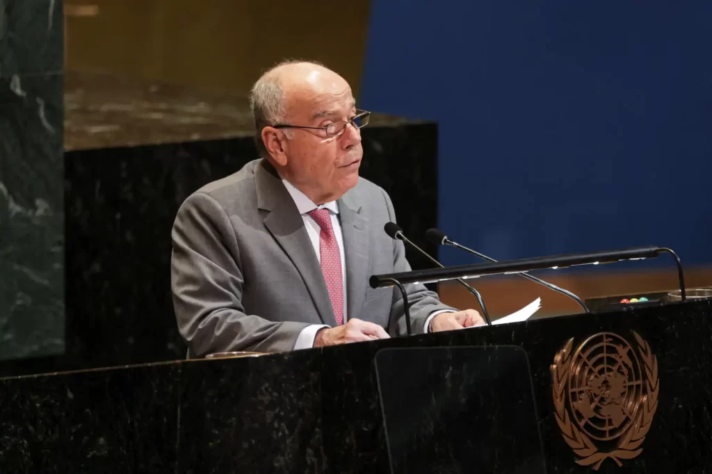 Pessoa idosa, homem de óculos, falando em um microfone durante um evento na ONU. Traje social Cinza e gravata rosa, fundo azul escuro.