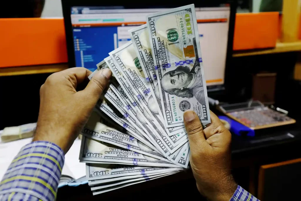 Pessoa segurando uma pilha de notas de dólar na frente de um computador, simbolizando dinheiro ou sucesso financeiro.
