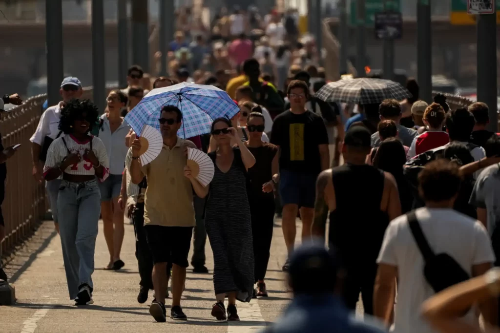 Pessoas caminhando na rua sob o sol forte, usando guarda-chuvas e roupas leves para se proteger do calor, em uma cena de aglomeração urbana