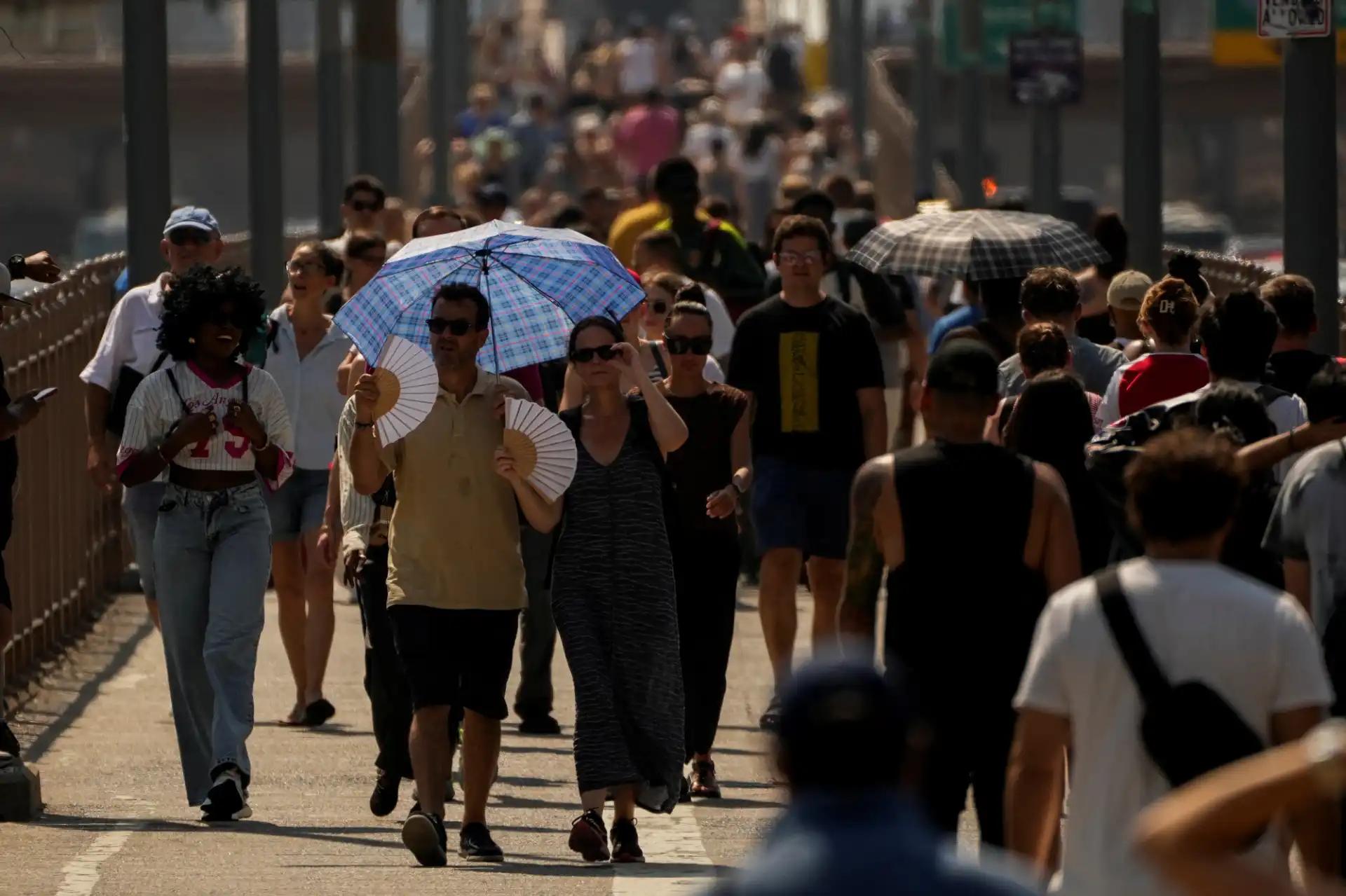 Pessoas caminhando na rua sob o sol forte, usando guarda-chuvas e roupas leves para se proteger do calor, em uma cena de aglomeração urbana