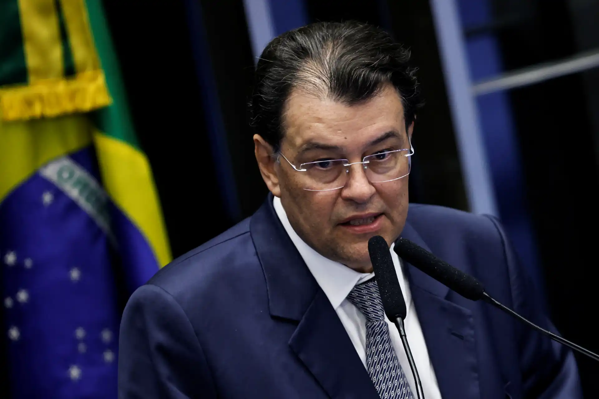 Foto de um político brasileiro falando em um evento oficial, vestindo terno e gravata, com Bandeira do Brasil ao fundo. Ideal para conteúdo relacionado à política brasileira.