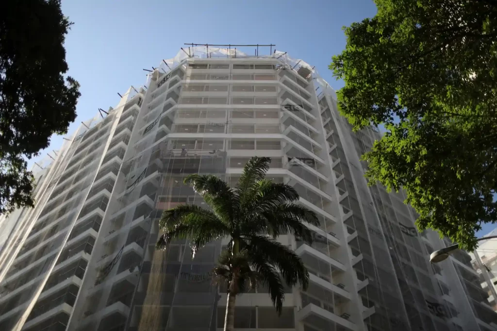 Imagem de um prédio em construção com estrutura de concreto, cercado por árvores e um céu claro ao fundo, destacando-se na paisagem urbana.