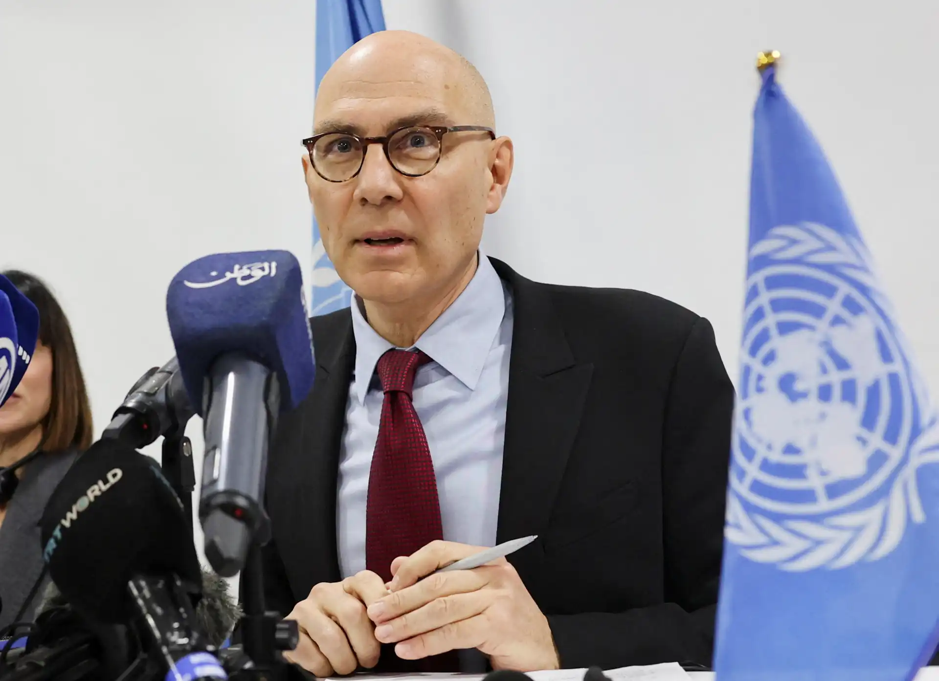 Imagem de um homem, provável representante ou porta-voz da ONU, durante uma entrevista de imprensa, com bandeiras da ONU ao fundo, vestindo terno e gravata vermelha.