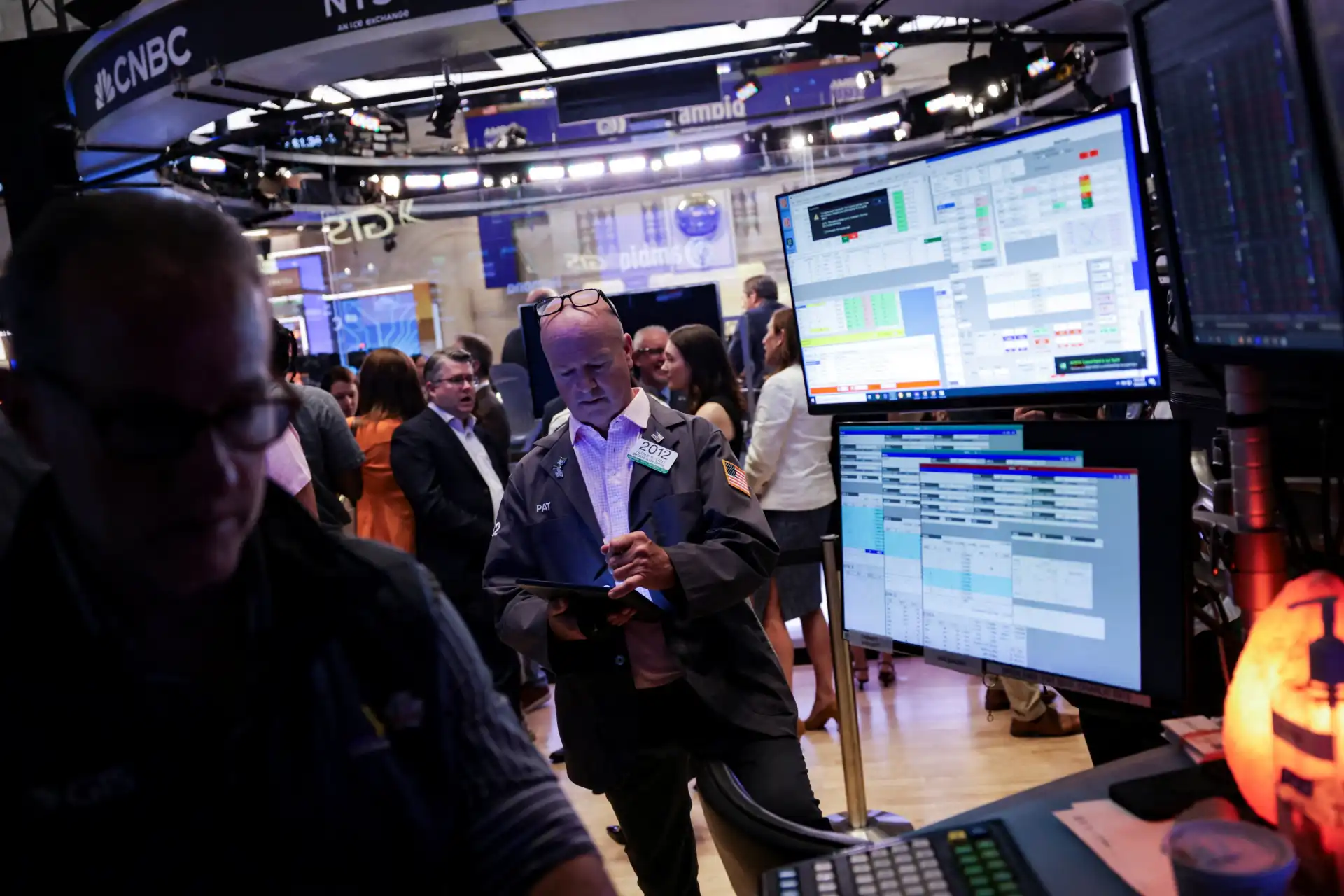 Imagem de uma sala de negociação na Bolsa de Valores, com várias telas exibindo gráficos e dados financeiros, pessoas analisando informações de mercado e operadores em ação.