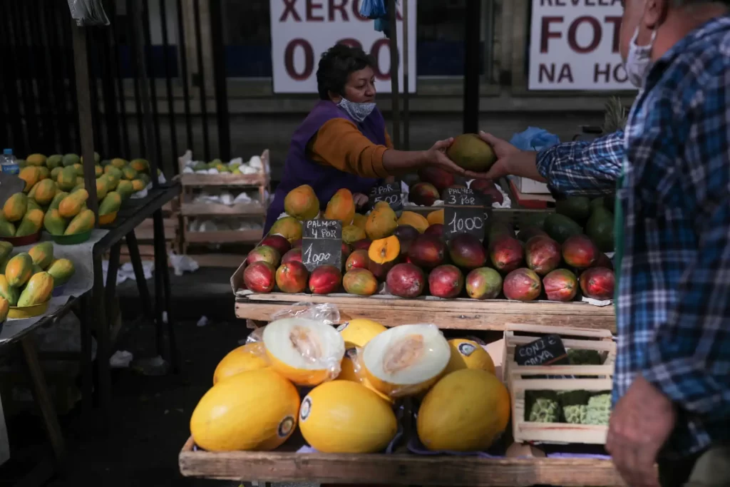 Mercado passa a ver inflação abaixo de 5% este ano pela 1ª vez desde janeiro, mostra Focus Venda de frutas variadas em feira ao ar livre com uma mulher atendendo cliente, destaque para mamão, maçã, manga e melões. Ambiente de feira com várias caixas de frutas frescas.
