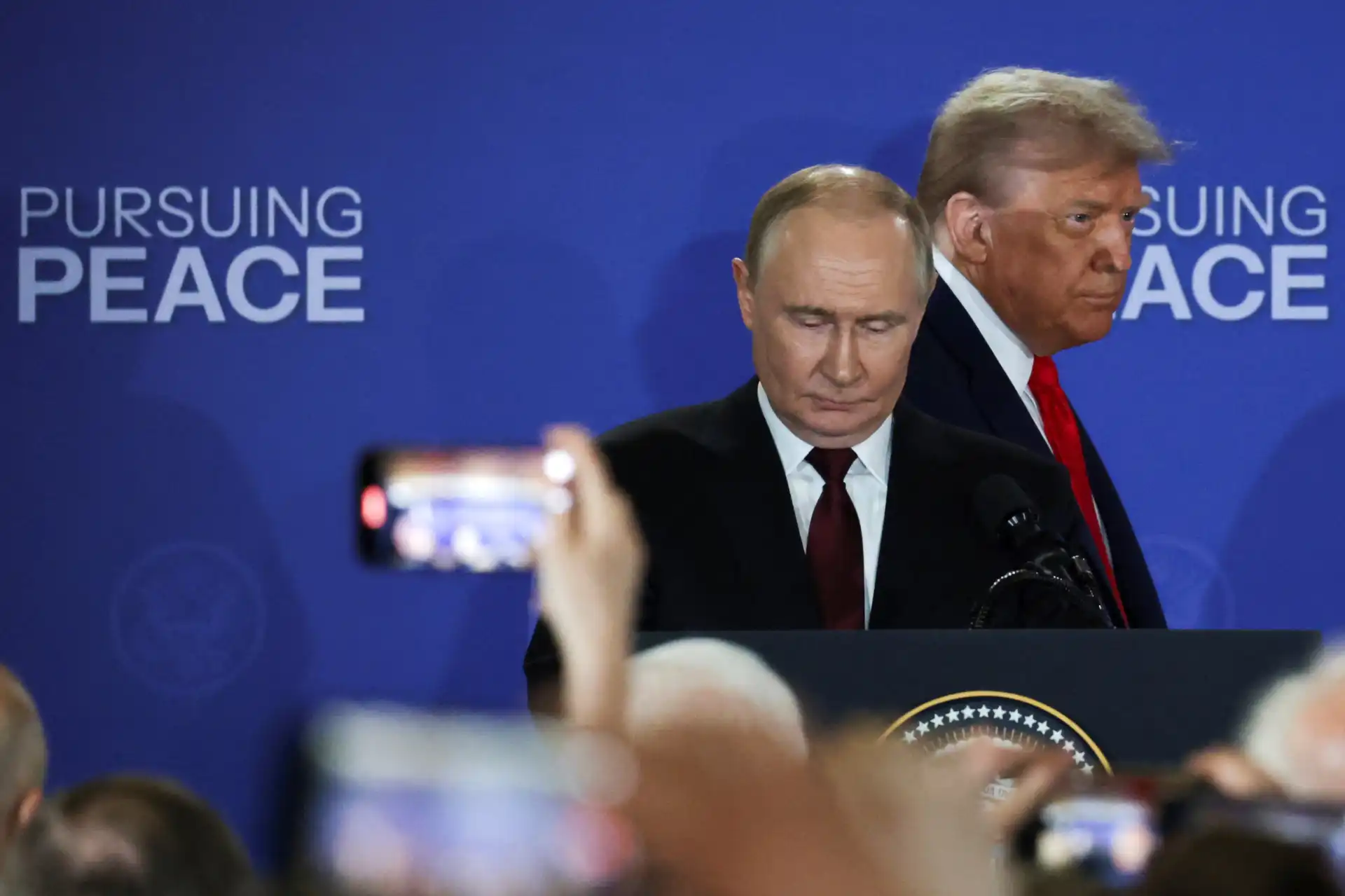 Imagem de Vladimir Putin e Donald Trump em evento de paz, com fundo azul e as palavras 'Pursuing Peace'.