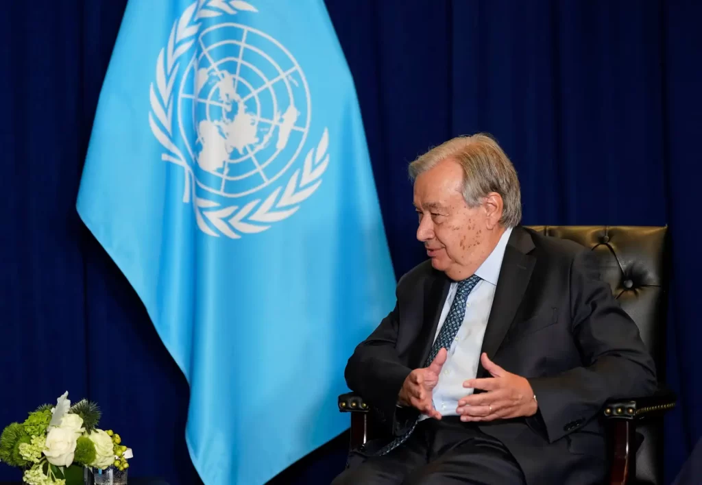 António Guterres, secretário-geral da ONU, em entrevista ou reunião, com a bandeira da Organização das Nações Unidas ao fundo. Ambiente formal, mostrando liderança e compromisso internacional.