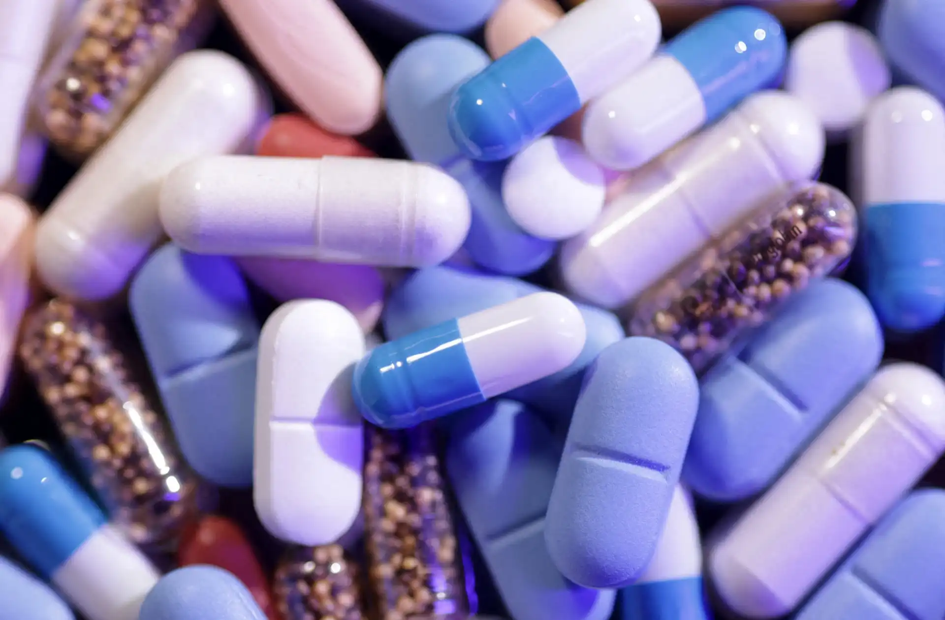 Imagem de cápsulas de medicamentos em cores azul, branco e roxo, incluindo algumas com bolinhas douradas, representando suplementos ou medicamentos com destaque para a palavra-chave
