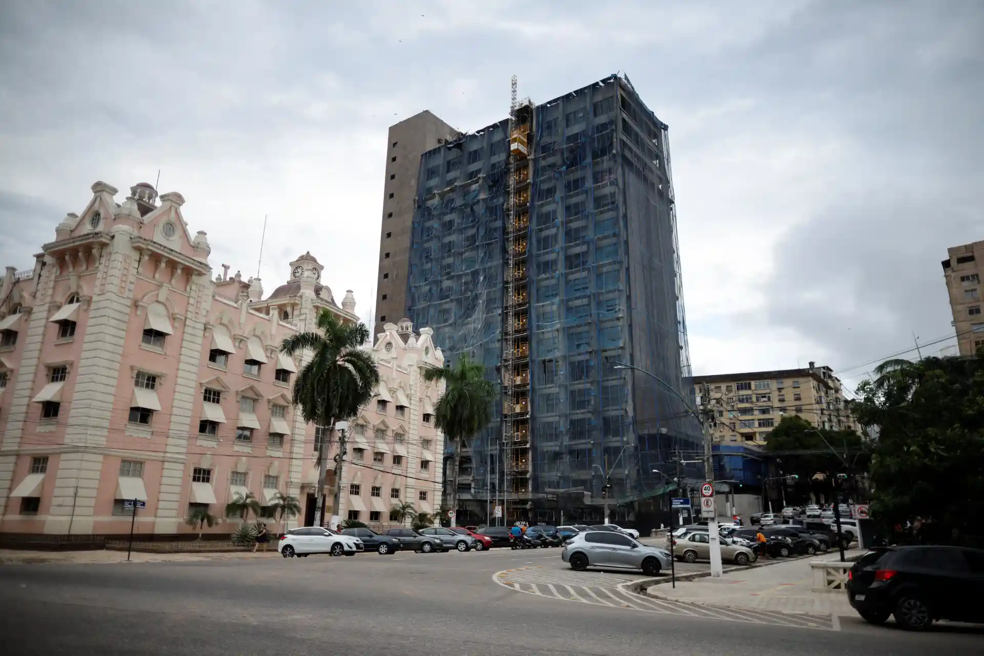 Construção de um prédio alto em uma cidade, com edifícios clássicos ao redor e árvores, sob céu nublado.
