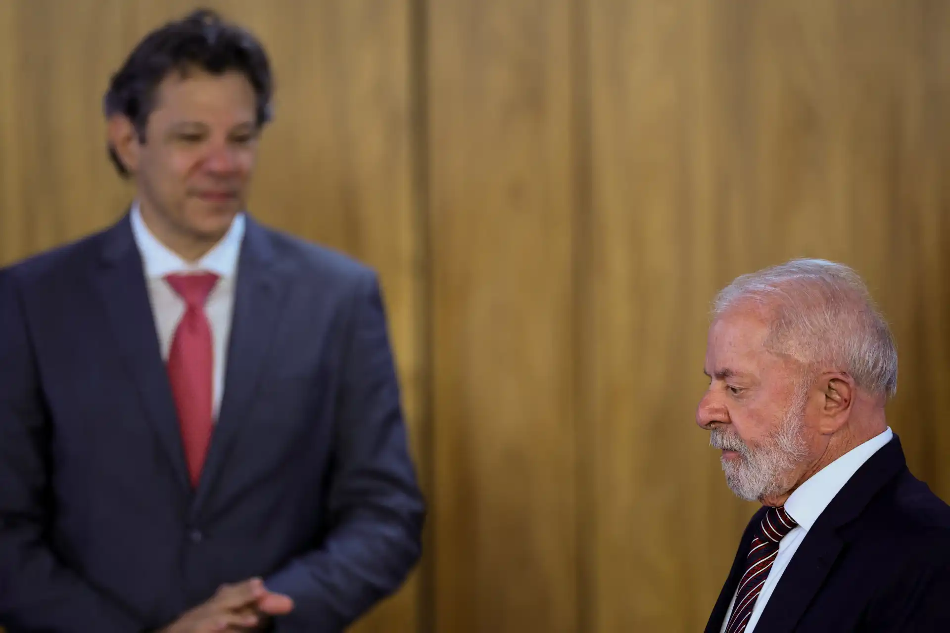 Homem mais jovem e homem mais velho em uma conversa formal, ambos usando ternos, com fundo de madeira, evidenciando um ambiente profissional.
