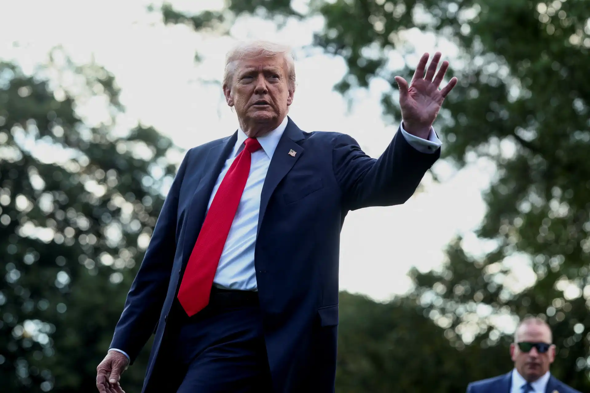 Imagem de Donald Trump ao ar livre, vestindo terno escuro, camisa branca e gravata vermelha, fazendo sinal de despedida ou cumprimento com a mão. Fundo com árvores e céu claro.