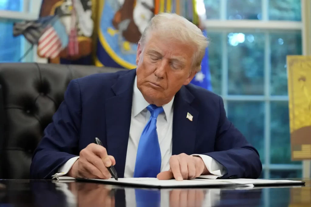 Imagem de Donald Trump, ex-presidente dos Estados Unidos, assinando documento no escritório da Casa Branca, com uma expressão concentrada.