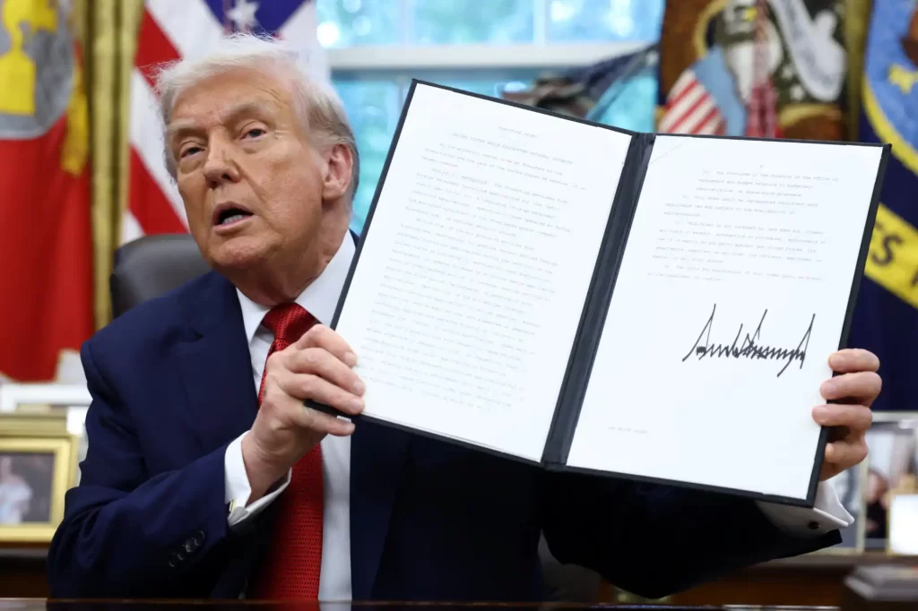 Imagem do ex-presidente dos Estados Unidos, Donald Trump, segurando uma cópia de um documento assinado, com uma assinatura e texto visíveis na página.