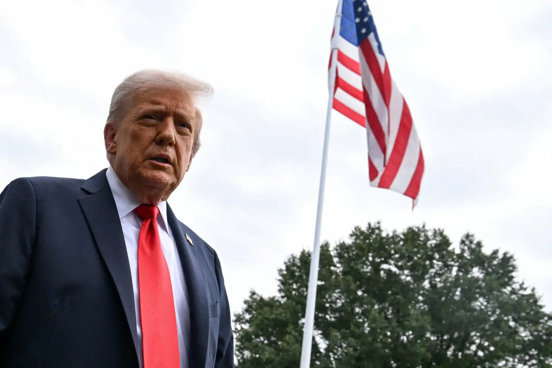 Imagem de Donald Trump ao ar livre durante um evento político, com bandeira dos Estados Unidos ao fundo, vestindo terno e gravata vermelha