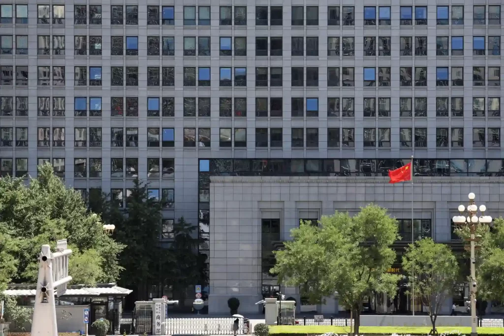 Imagem do edifício de um órgão governamental na China, com bandeira chinesa hasteada, árvores na frente e janelas refletindo o ambiente externo.