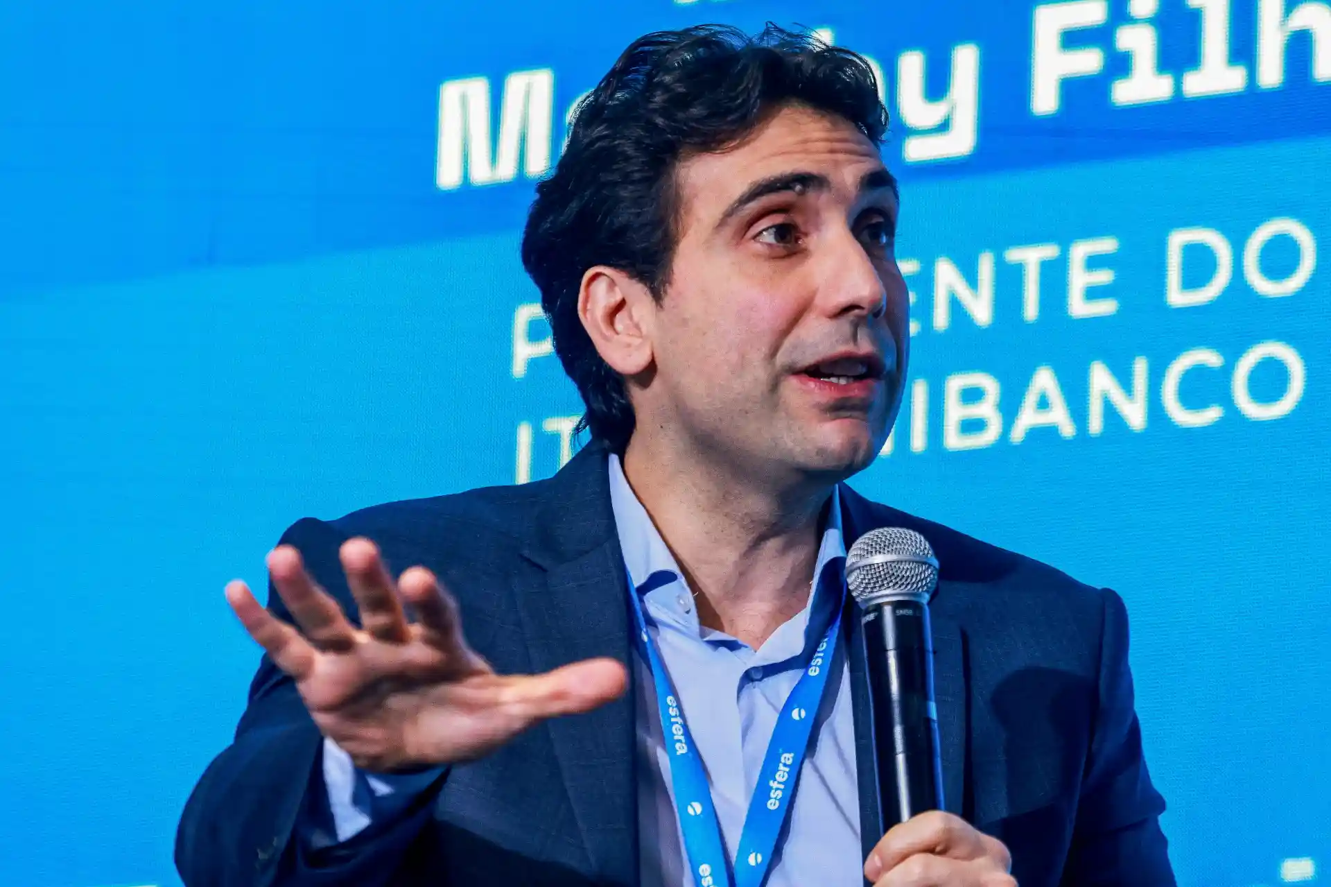 Executivo ou palestrante falando em evento do Banco Itaú, com fundo azul e frase de destaque, usando blazer e camisa, segurando microfone.