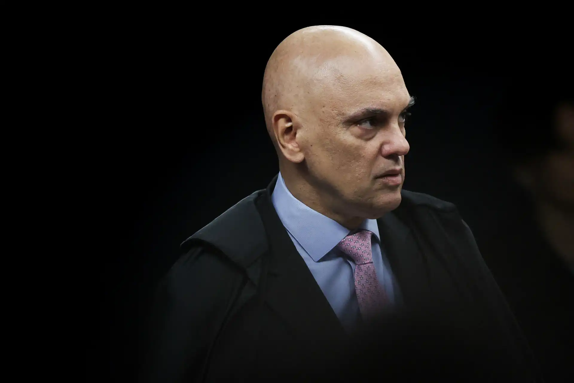 Imagem de um homem com cabeça calva, expressão séria, vestindo terno com camisa azul e gravata rosa escuro, em fundo preto.