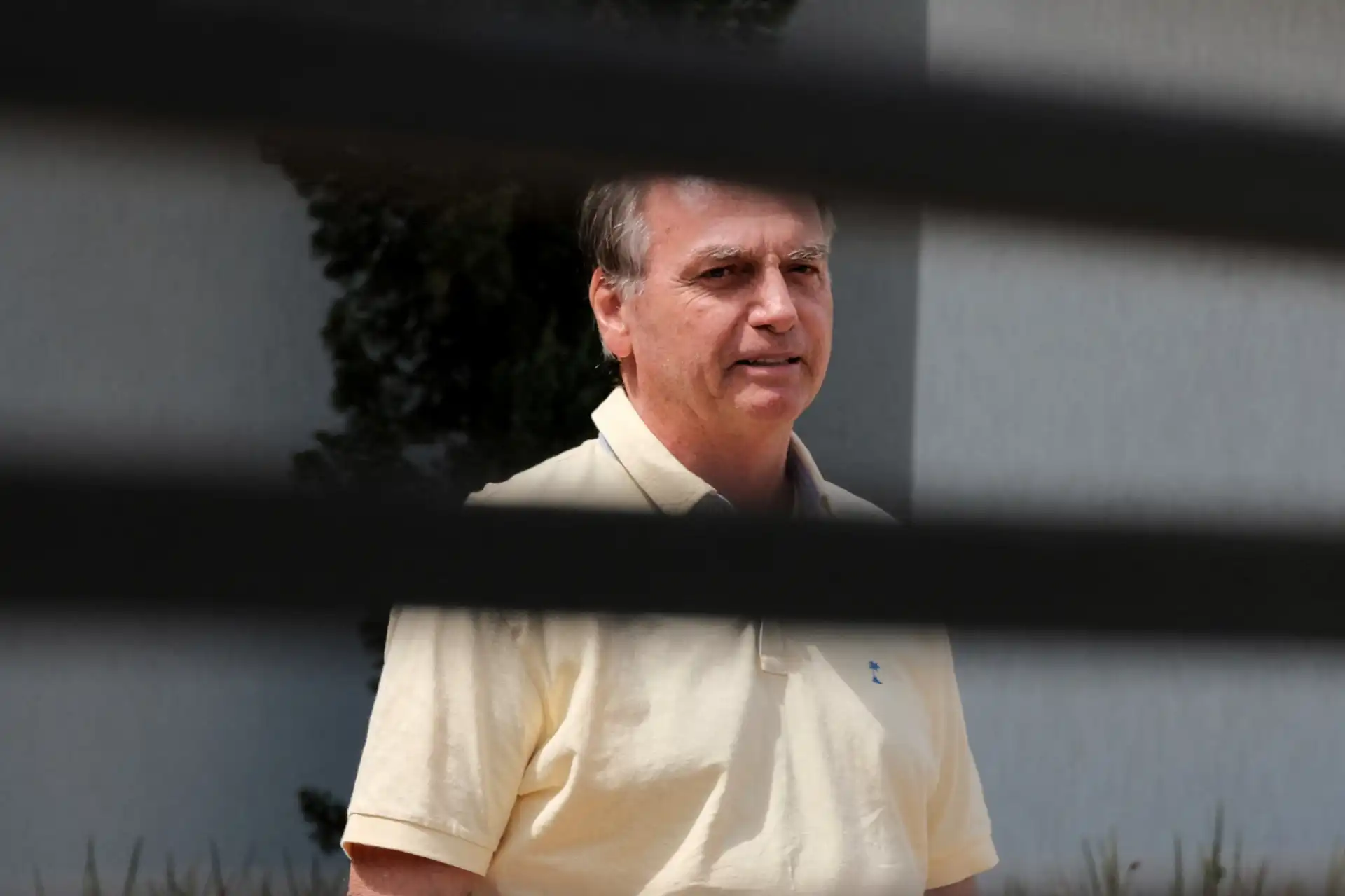 Foto de um homem de cabelo castanho claro, usando uma camisa amarela clara, em ambiente externo durante o dia, visto através de uma cerca ou grades.
