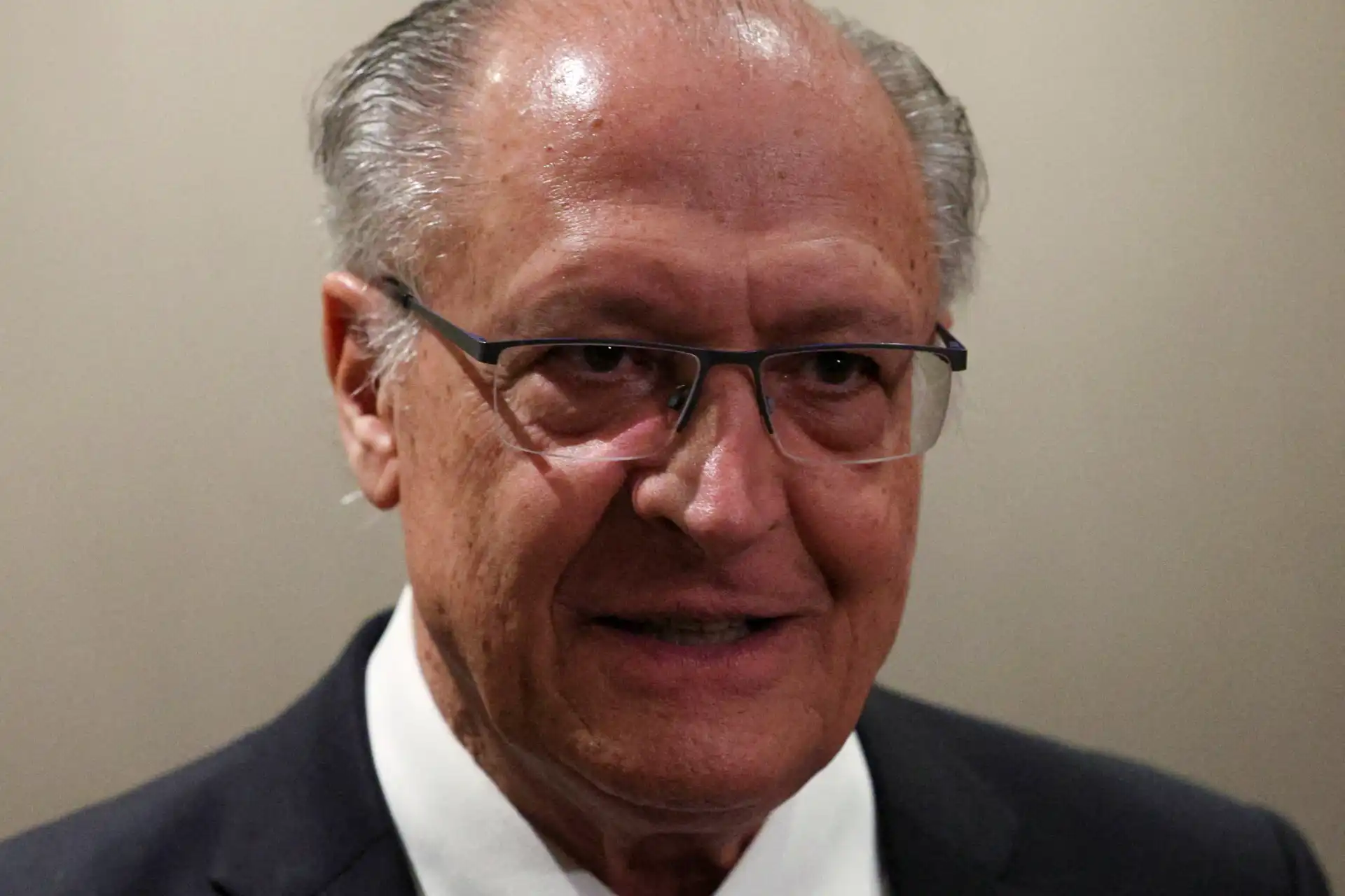 Homem de aparência idosa com cabelo grisalho, usando óculos e terno, sorrindo para a câmera, em ambiente neutro, ideal para conteúdo sobre envelhecimento saudável.