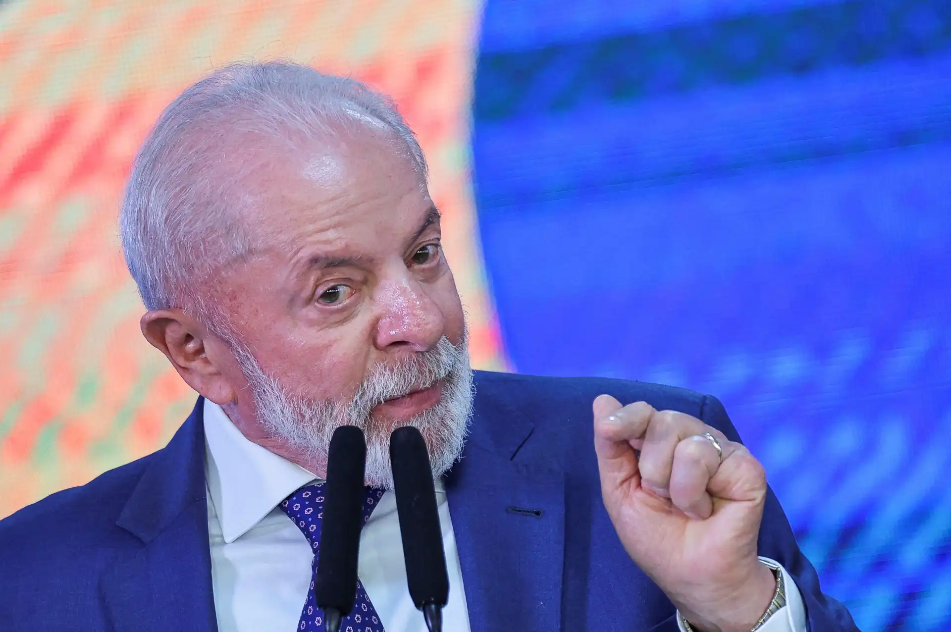 Homem idoso com barba branca e cabelo grisalho, usando terno azul escuro e gravata azul, fazendo um gesto de afirmação durante uma fala em público.