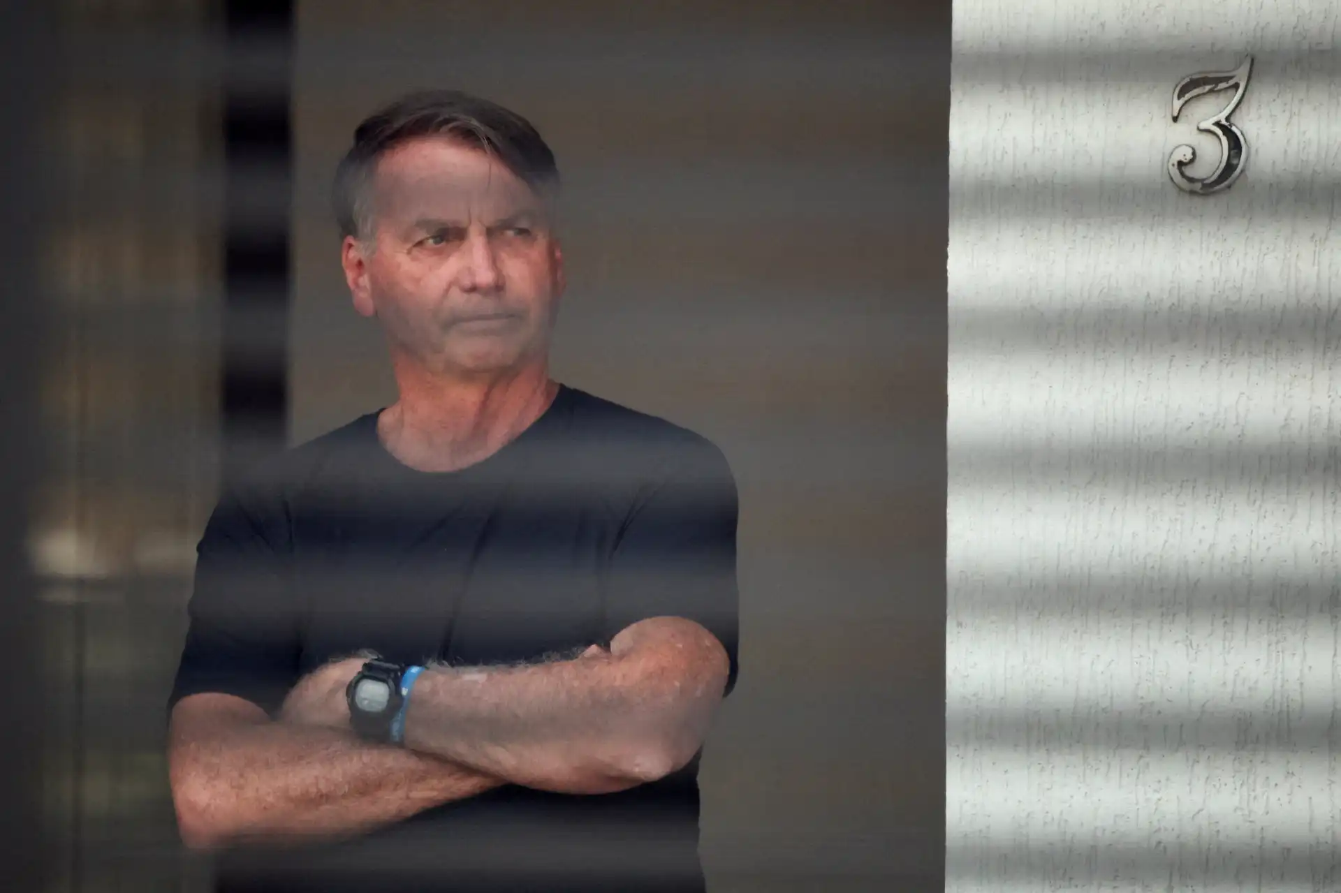 Homem pensativo observando ao lado enquanto está atrás de uma janela com reflexos, vestindo uma camiseta preta e usando relógio no pulso.