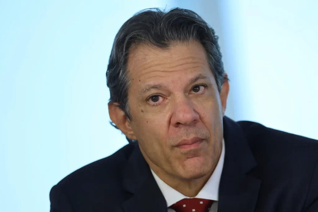 Homem de terno azul escuro e gravata vermelha com pontos brancos, expressão séria, cabelo castanho com fios grisalhos, em ambiente de escritório.