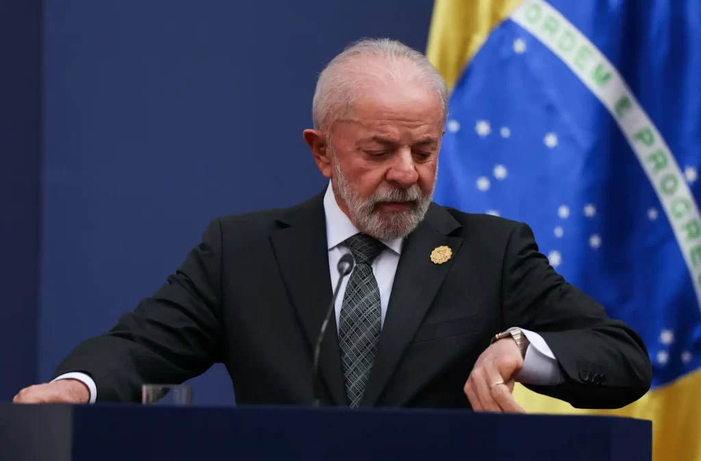 Lula, presidente do Brasil, olhando para o relógio durante uma coletiva de imprensa, com bandeira brasileira ao fundo.