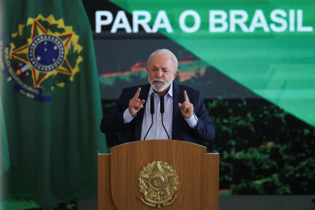 Luiz Inácio Lula da Silva durante discurso oficial no Brasil, na presença da bandeira nacional, usando terno, com expressão séria e gesticulando com as mãos, destacando a importância da política brasileira.