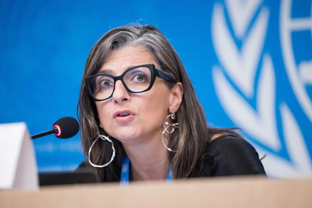 Mulher falando em uma conferência, com o logo da ONU ao fundo, usando óculos grandes e brincos grandes, representando uma figura de autoridade na ONU.