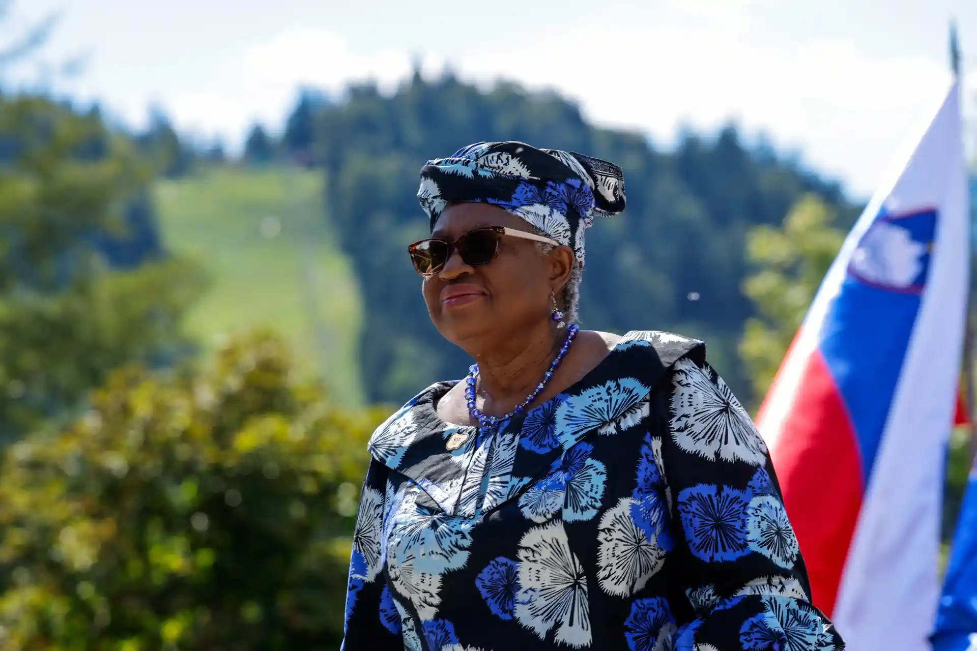 Mulher idosa usando chapéu, óculos escuros e roupa azul com estampa de flores, ao ar livre com bandeira da Eslováquia ao fundo, simbolizando orgulho nacional e identidade cultural.