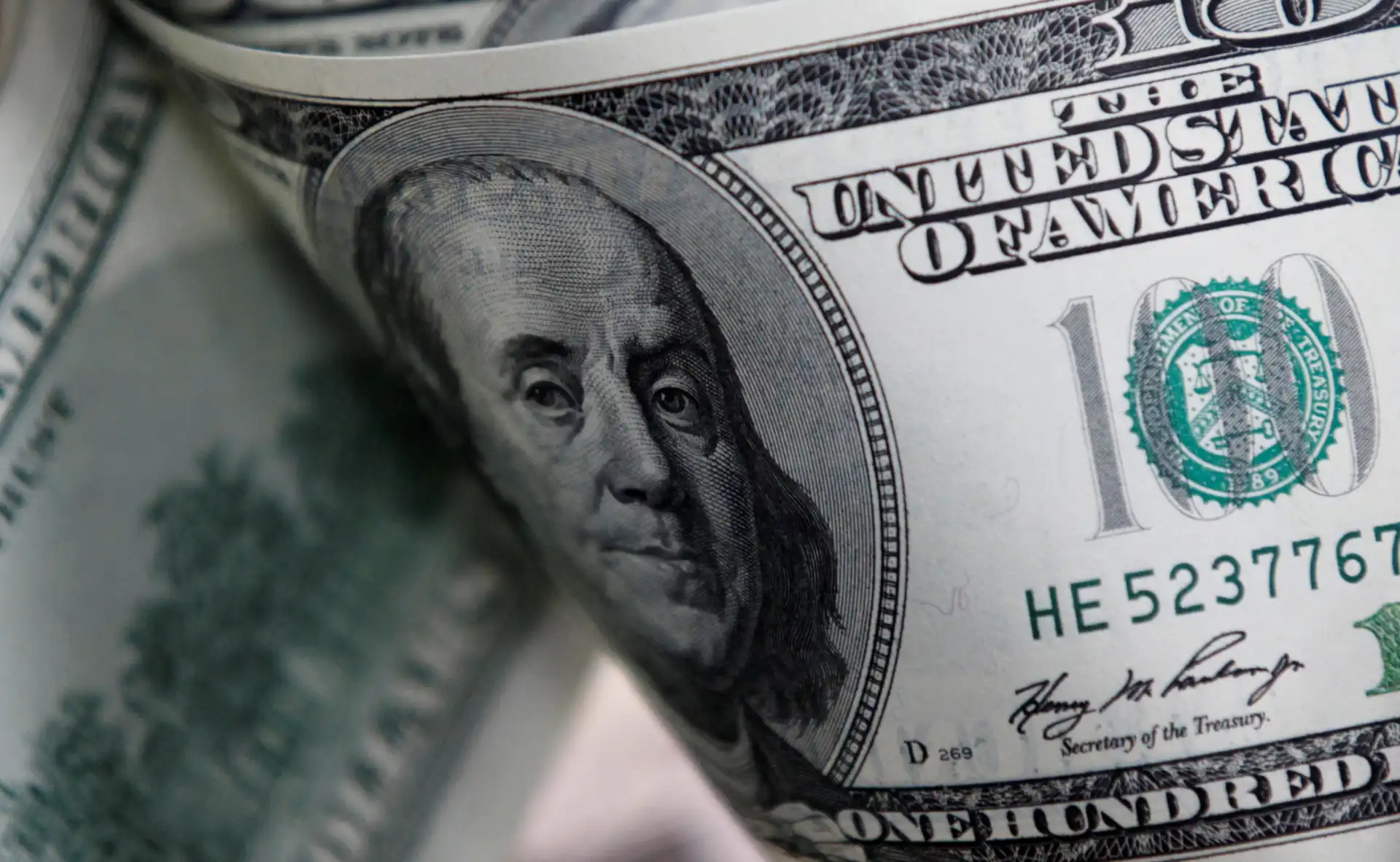 Imagem de uma nota de cem dólares americanos com o rosto de Benjamin Franklin em foco e detalhes visíveis da cédula