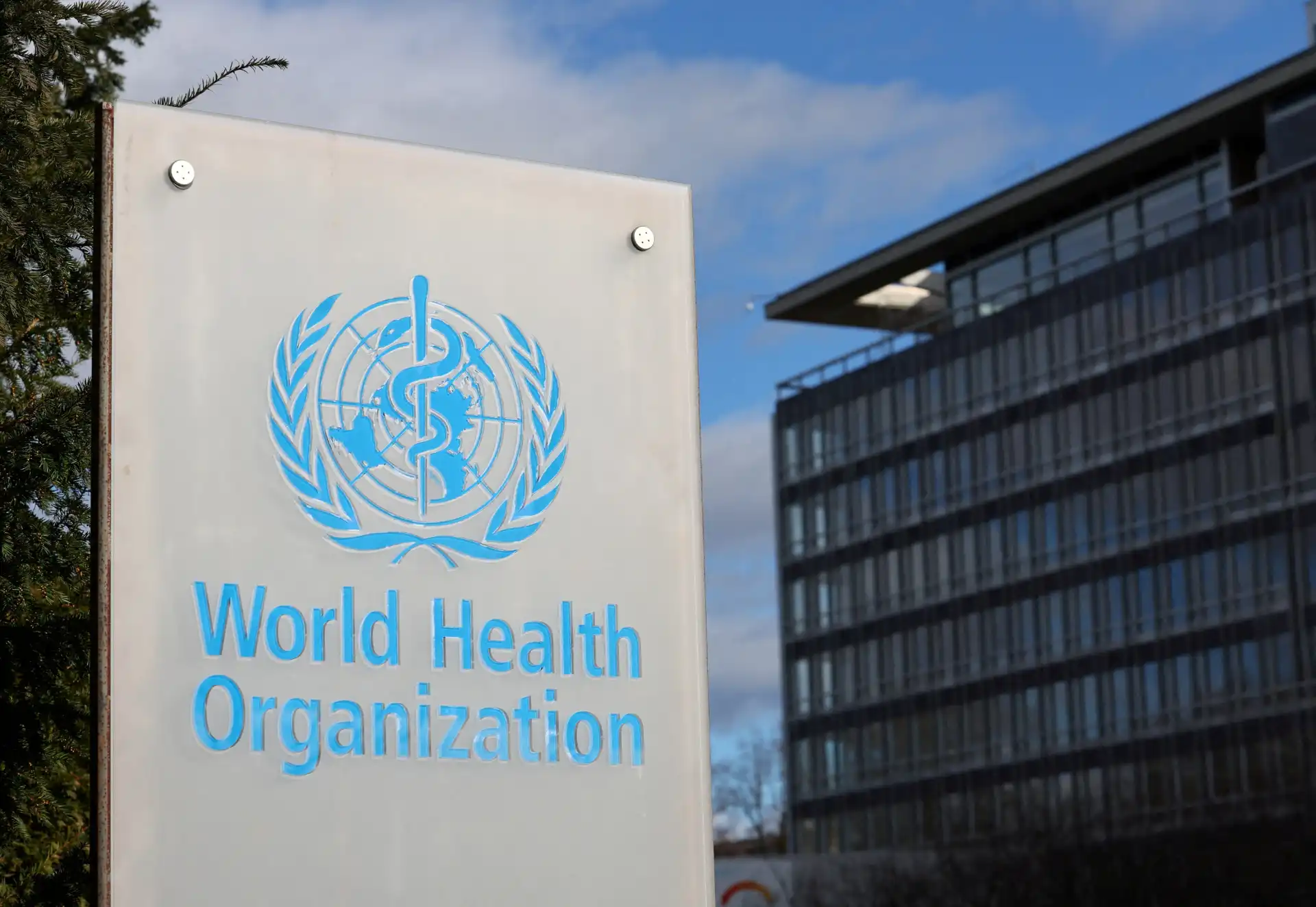 Imagem do prédio da Organização Mundial da Saúde (OMS) em um dia de céu azul, com destaque para o logotipo da OMS na placa de sinalização.