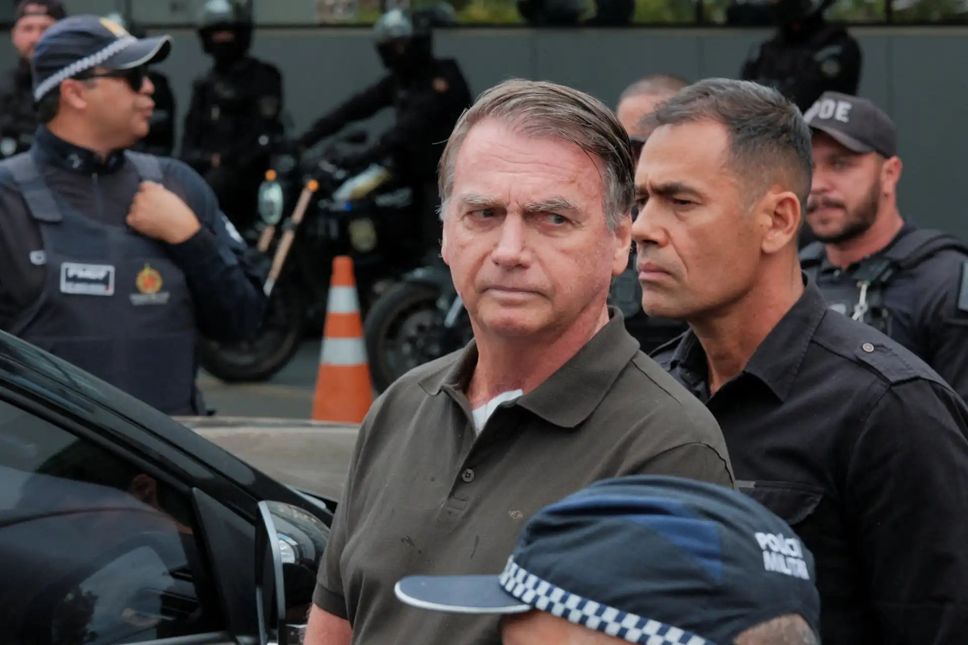 Imagem de um político brasileiro em uma manifestação com presença policial e pessoas ao fundo.
